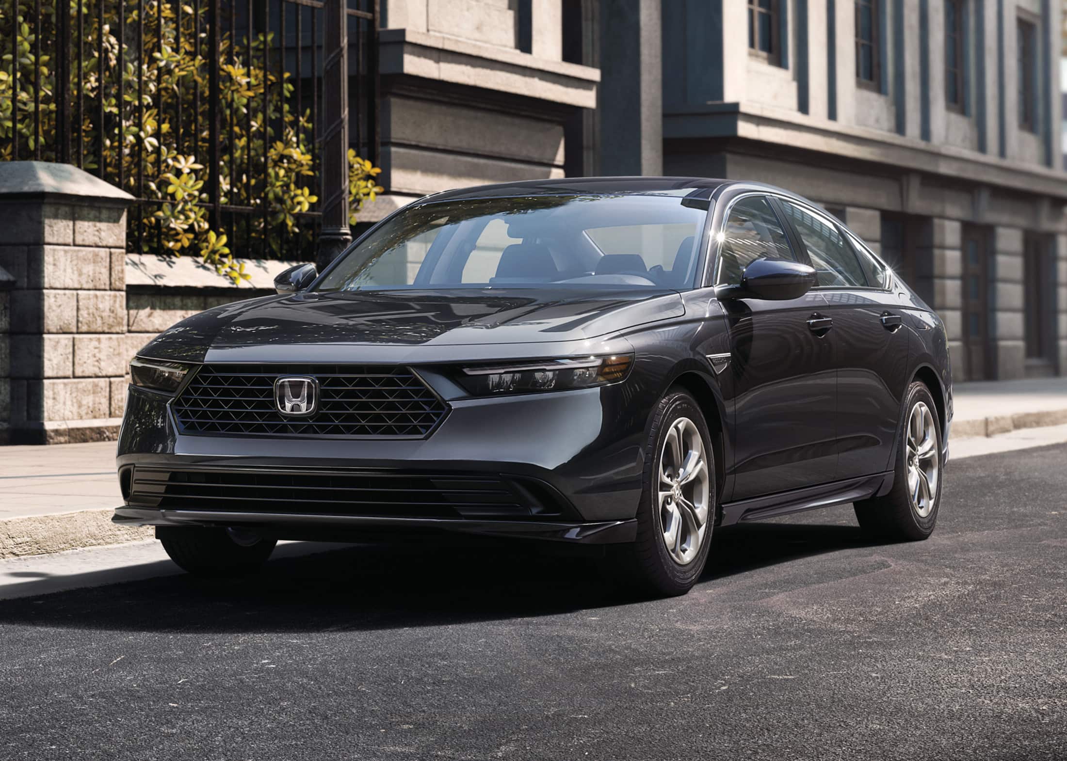 7/8 front driver-side view of the 2023 Honda Accord LX in Meteorite Gray Metallic, shown parked on the street next to a building and a city park with trees.