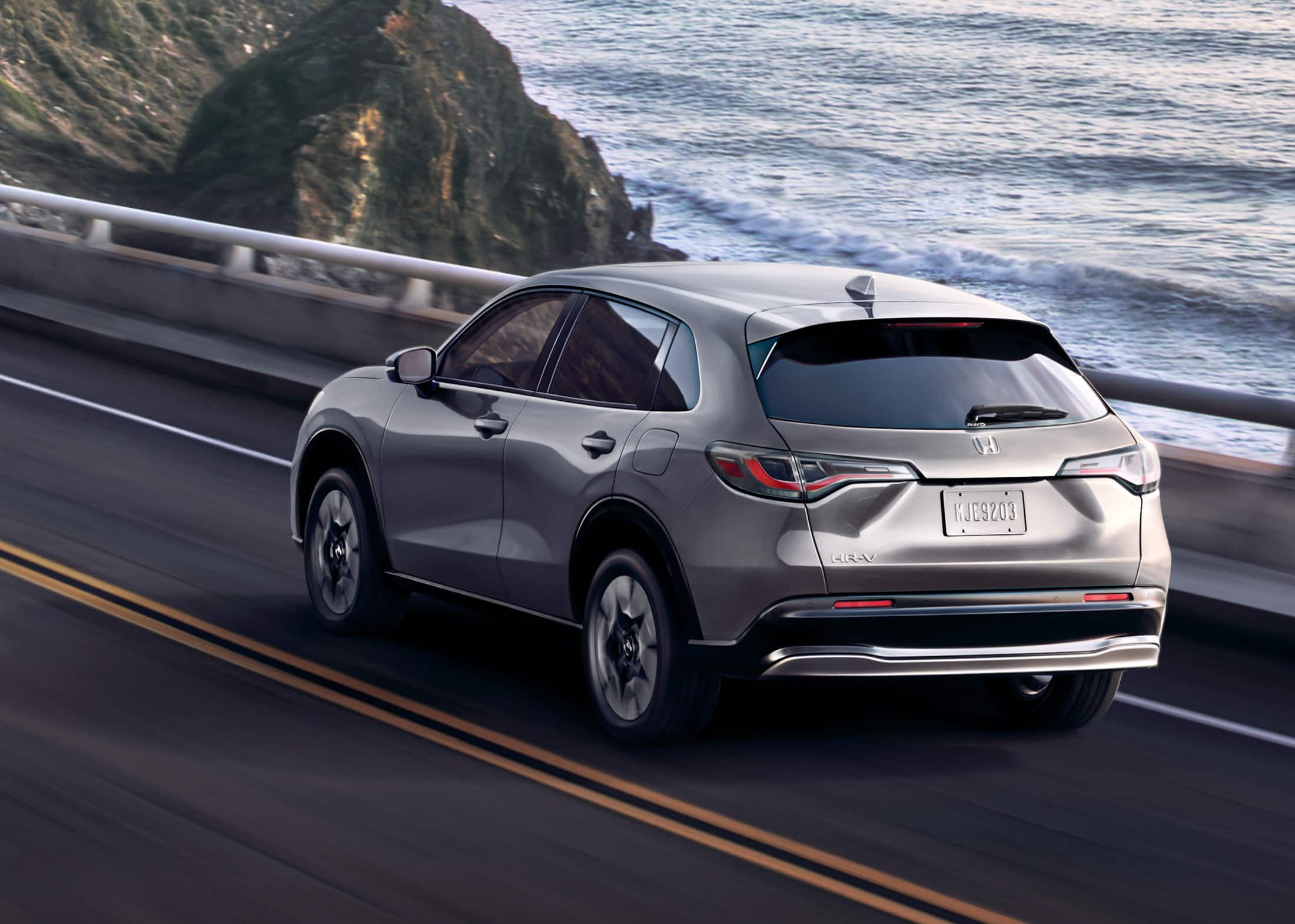 7/8 rear driver-side view of the 2026 Honda HR-V EX-L in Modern Steel Metallic, shown driving along a coastal highway.