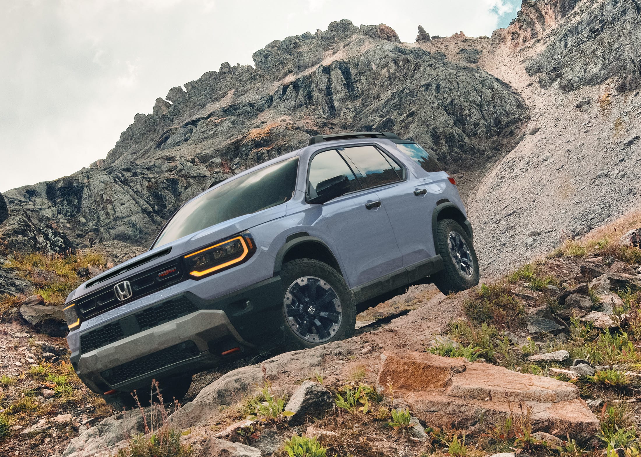 3/4 front driver-side view of the 2026 Honda Passport TrailSport Elite in Sonic Gray Pearl, shown descending down a rocky mountain path, with a black-spired mountaintop in background.